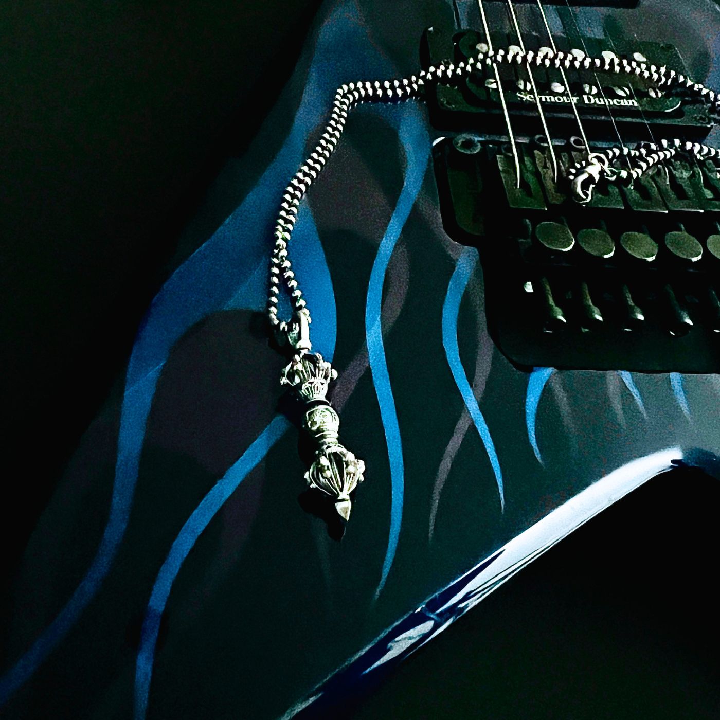 The WARBORN RELIC sterling silver pendant necklace displayed on a Jackson King V electric guitar with blue ghost flames in a backstage setting.