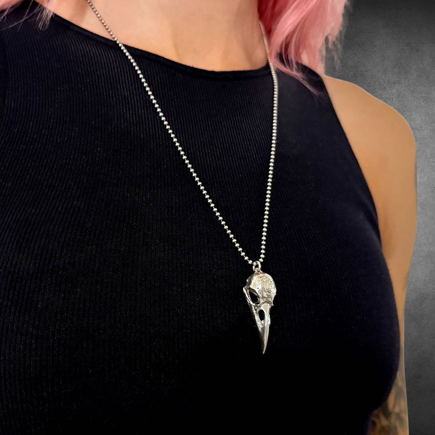 Person wearing a black top with a silver necklace featuring a bird skull pendant on a 24-inch ball chain. Otherwise known as CROW'D CONTROL
