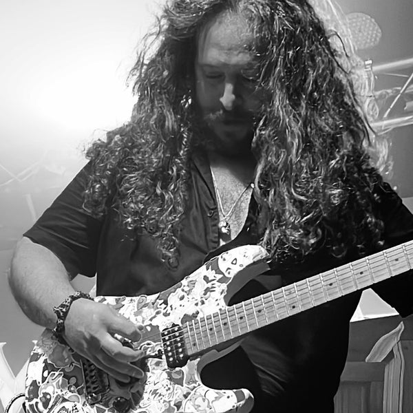 Alex 5000 wearing the FINAL LAUGH sterling silver skull pendant necklace by ROCKSTAR★REFINEMENTS™ on stage playing electric guitar. Black-and-white photograph of the long-haired guitarist performing.