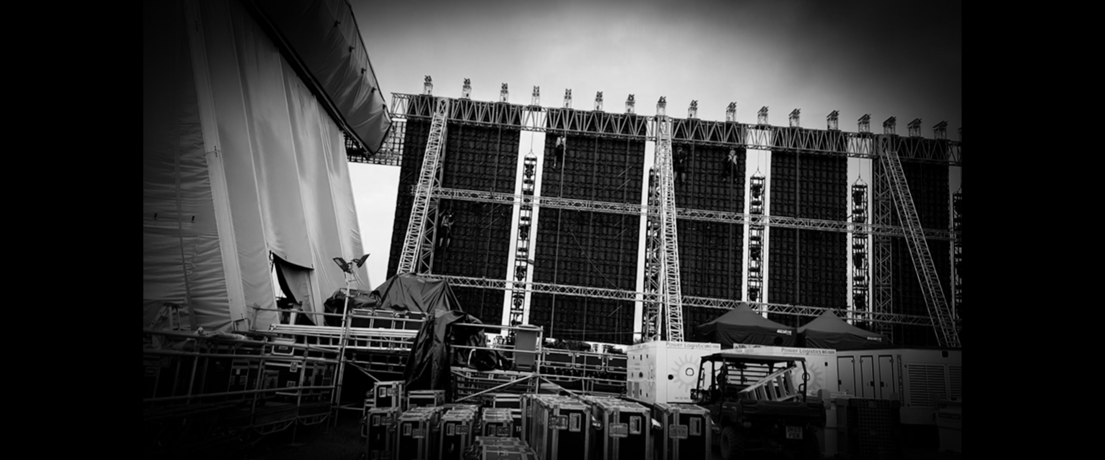 Backstage area of a large outdoor festival main stage in black and white, showing scaffolding, lighting rigs and flight cases.