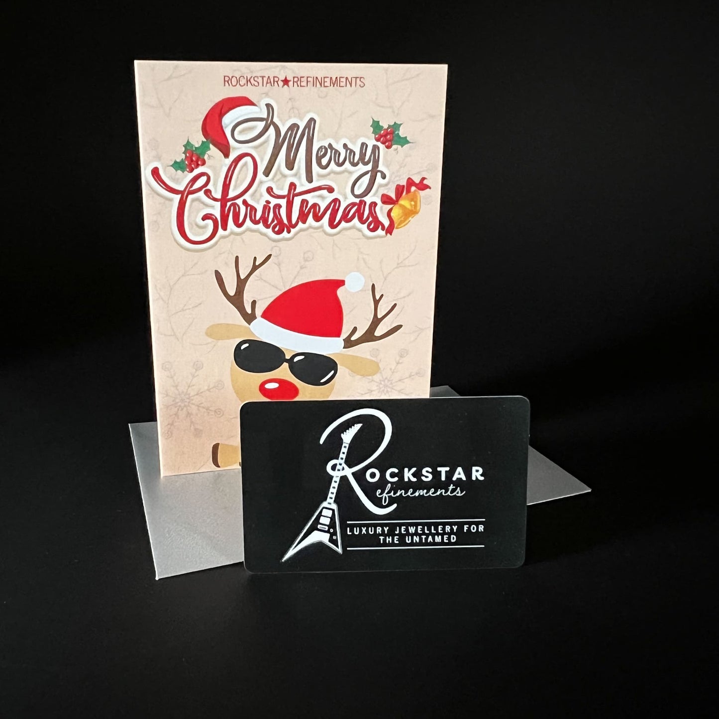 Rockstar reindeer Christmas card and silver envelope with a physical ROCKSTAR★REFINEMENTS gift voucher card photographed on a black background.