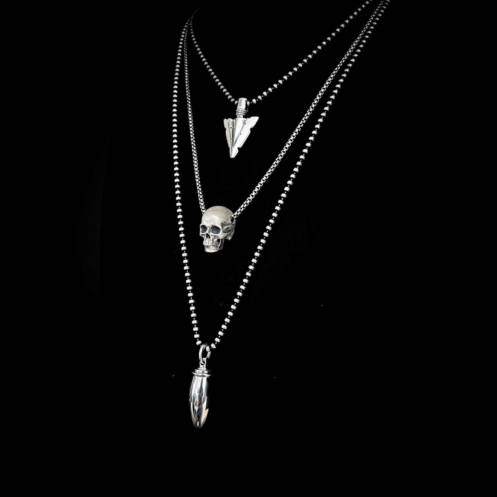 DEADLY LITTLE DEATAILS collection featuring FINAL POINT arrowhead, RESTING DEAD FACE skull and PRETTY LOADED pendants in sterling silver, on a black background.