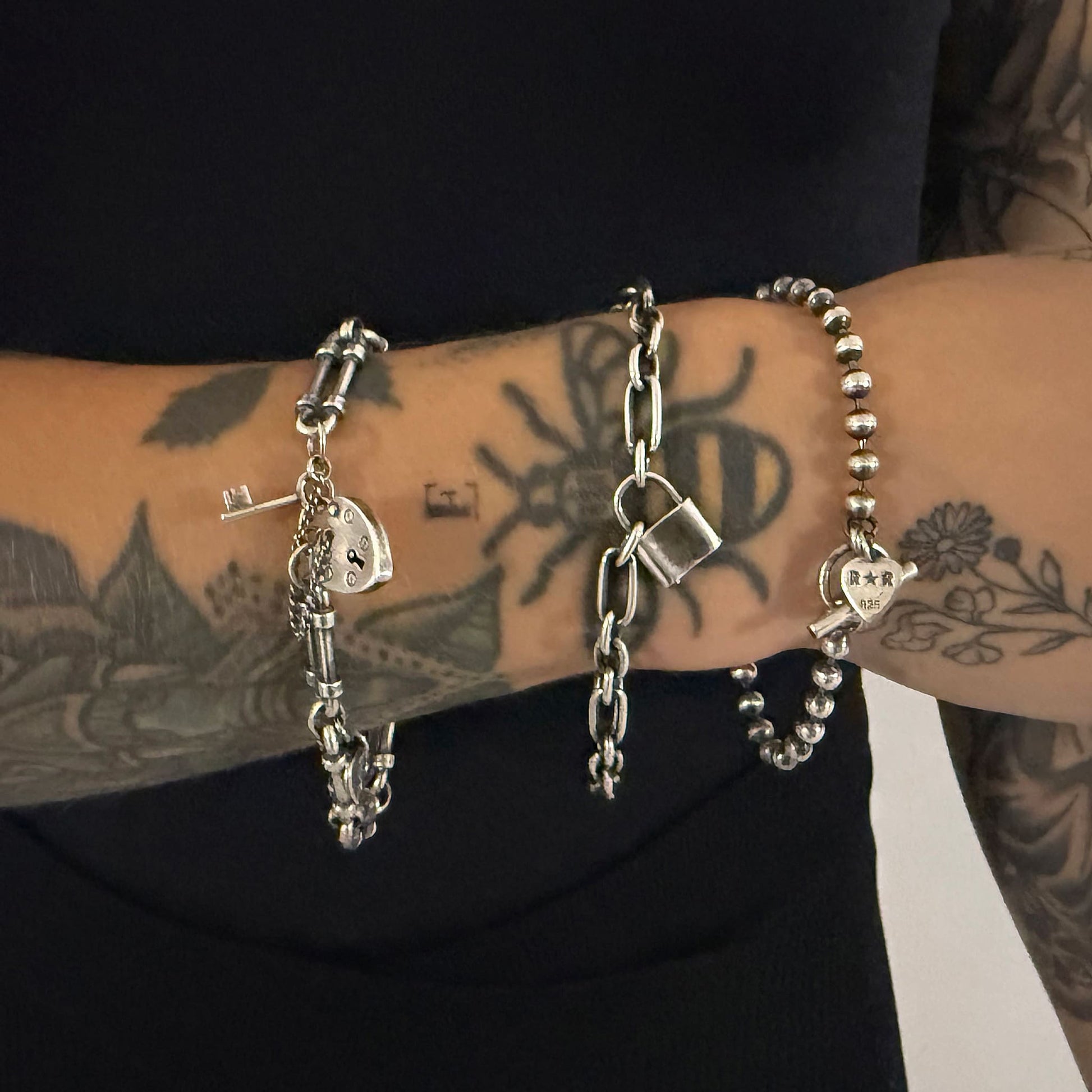Three sterling silver bracelets worn on a tattooed wrist — WRISTRICTED ACCESS on the left, LOCKED-IN WRIST CHAIN in the centre, and SIDEKICK SLAYER on the right.