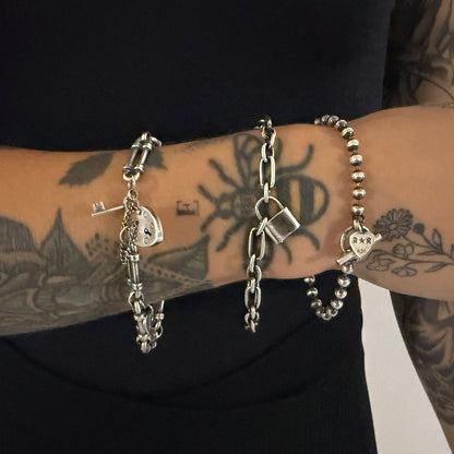 Three sterling silver bracelets worn on a tattooed wrist — WRISTRICTED ACCESS on the left, LOCKED-IN WRIST CHAIN in the centre, and SIDEKICK SLAYER on the right.