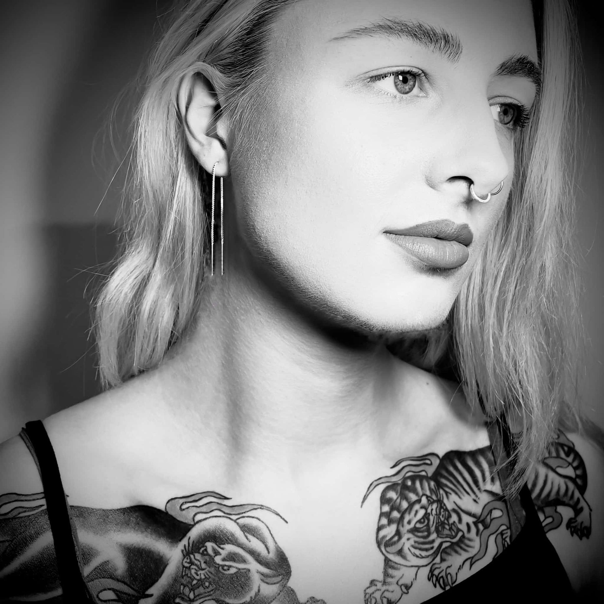 Black-and-white portrait of model wearing sterling silver Out of Line earrings by ROCKSTAR★REFINEMENTS™ with tattoo and piercing details visible.