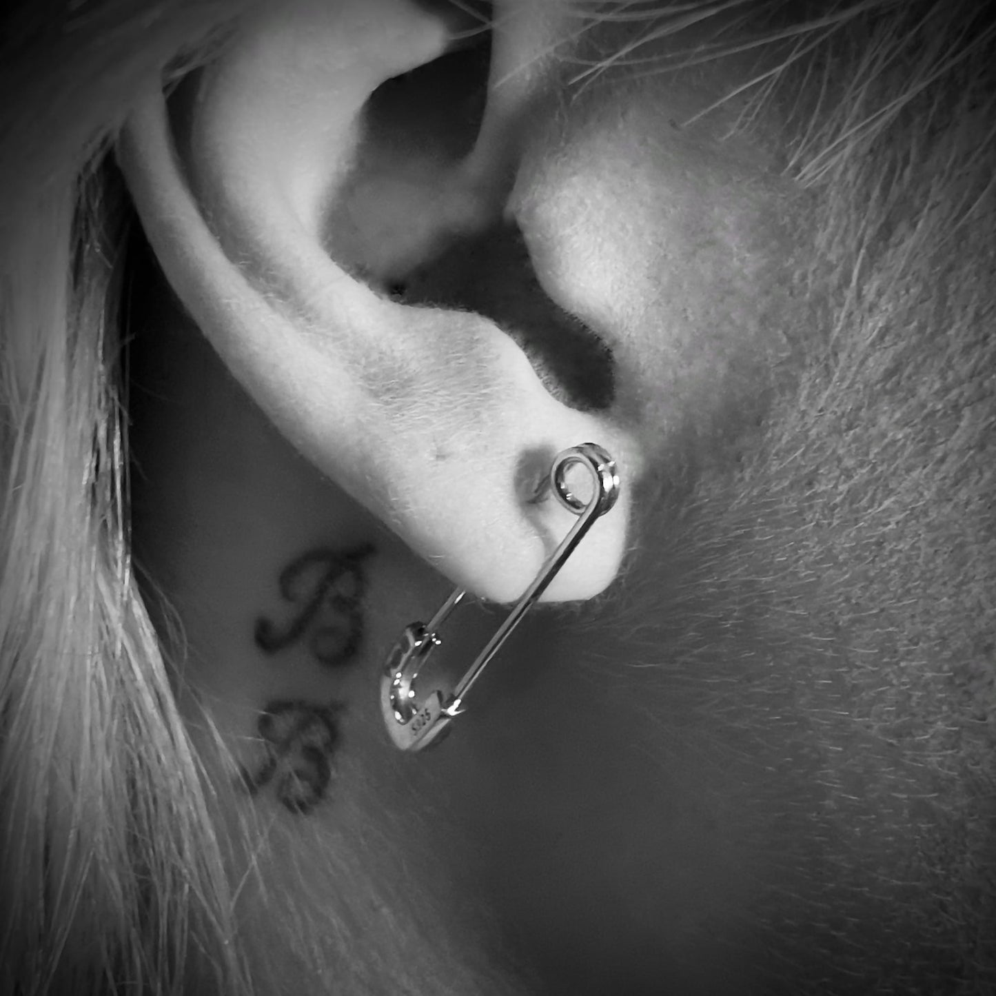 Close-up black-and-white photo of sterling silver SAFETY LAST safety pin earrings by ROCKSTAR★REFINEMENTS™ worn on ear with tattoo detail visible. 