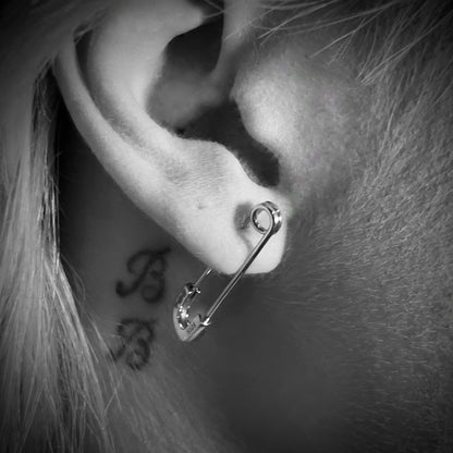 Close-up black-and-white photo of sterling silver SAFETY LAST safety pin earrings by ROCKSTAR★REFINEMENTS™ worn on ear with tattoo detail visible. 